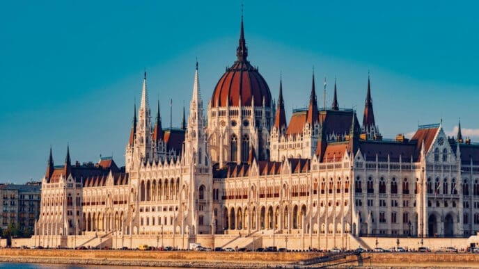 A full view of the Hungarian Parliament Building, a large Gothic Revival structure with a central dome and many spires, standing on the bank of a river under a clear blue sky.