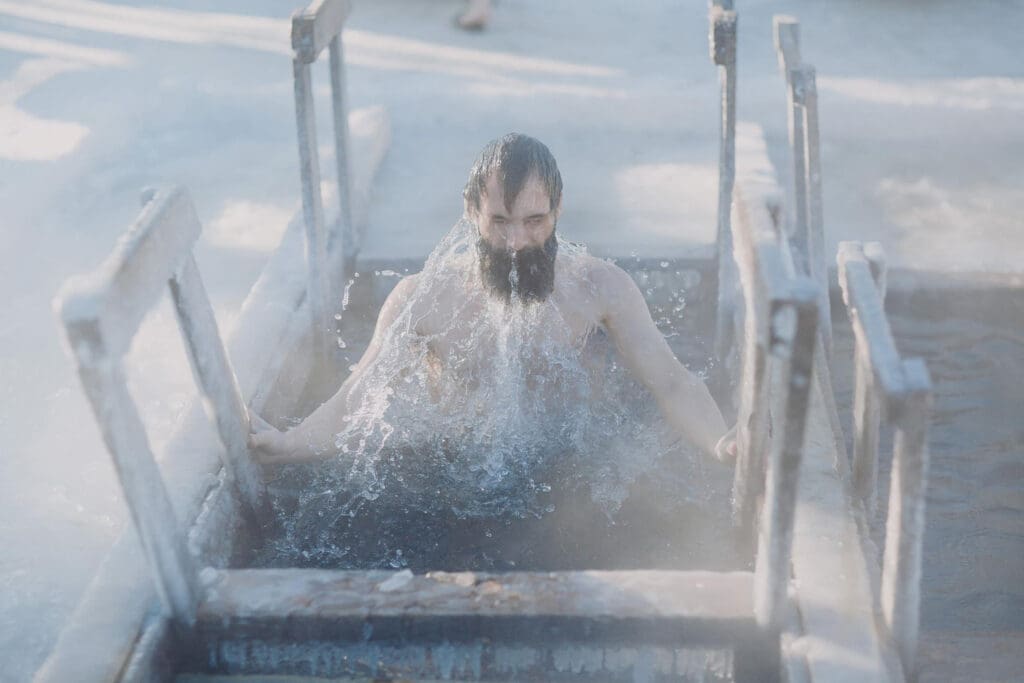 A bearded man emerges from a hole in the ice, splashing water as he climbs out.