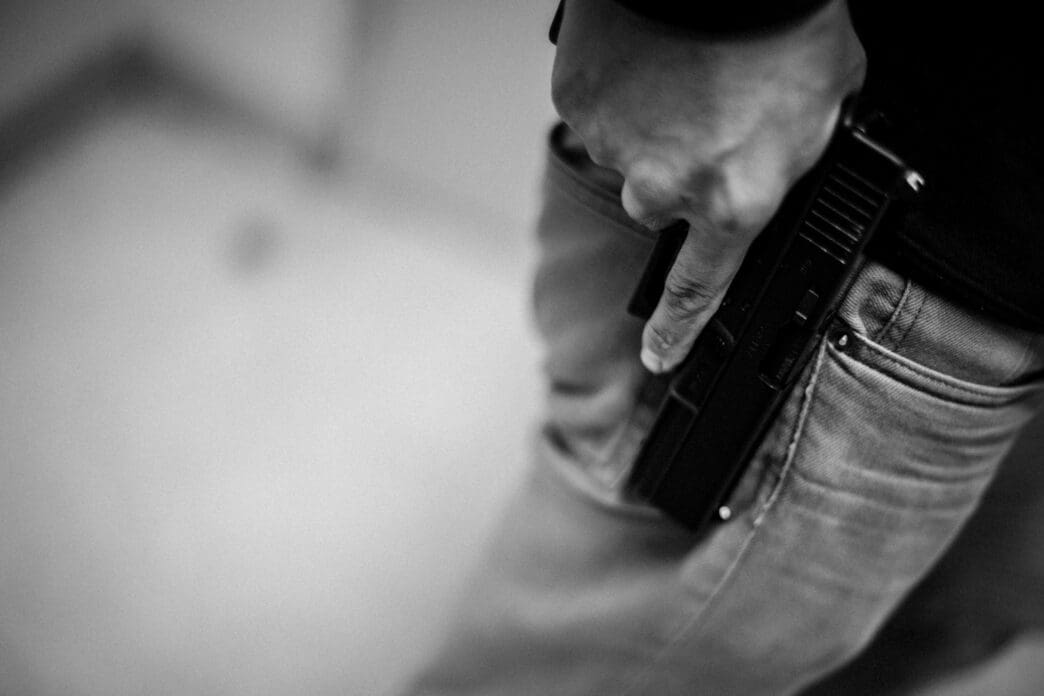A black and white photograph showing a person's hand gripping the handle of a handgun that is tucked into the back pocket of their jeans.