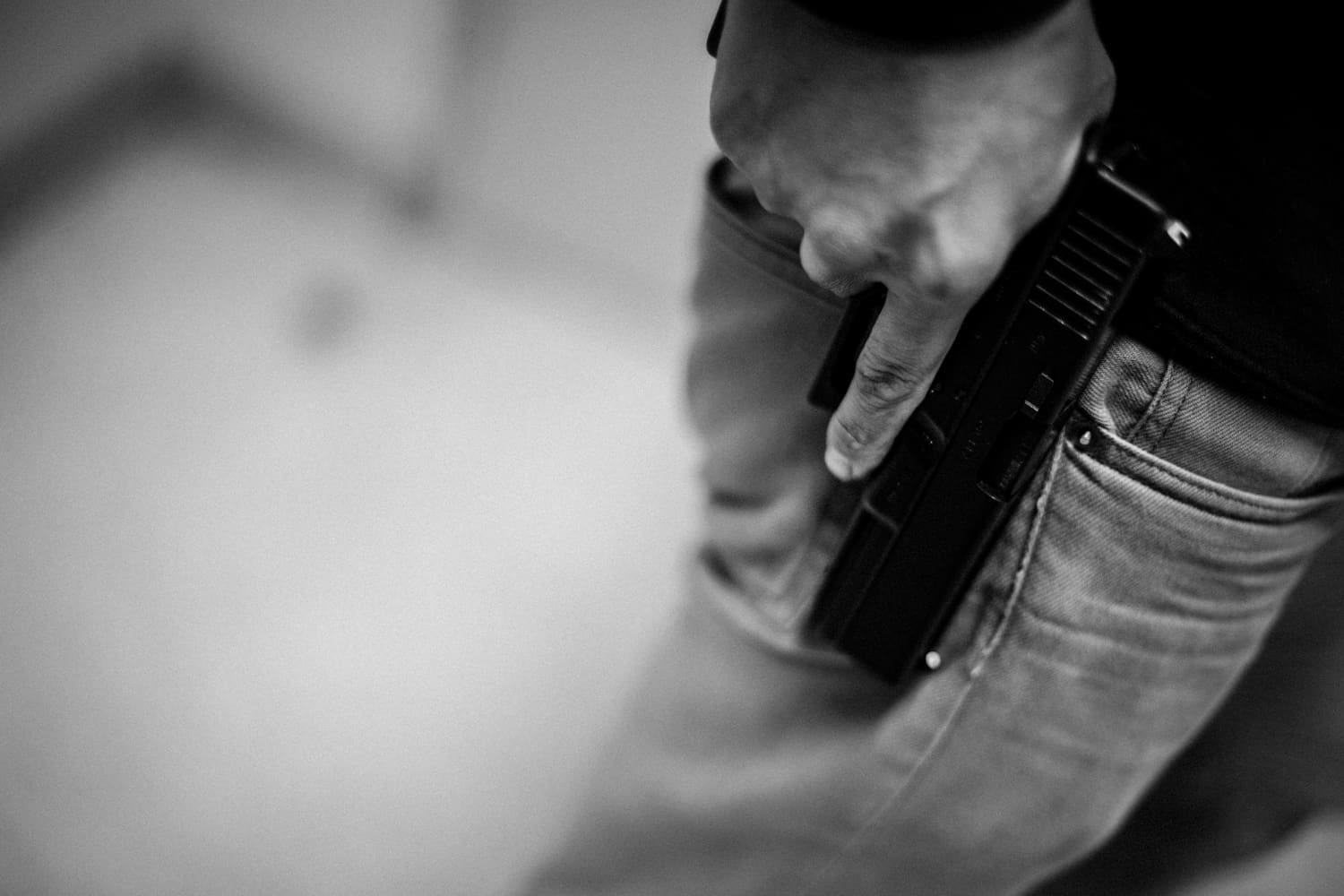 A black and white photograph showing a person's hand gripping the handle of a handgun that is tucked into the back pocket of their jeans.
