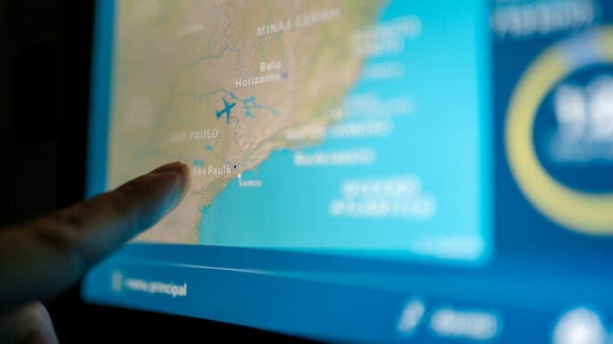 A passenger's finger touches an LCD entertainment screen on a plane, which displays a map of Brazil with an icon of a plane over the coast near São Paulo.