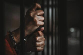 A close-up, dark photograph of a person's hands gripping the metal bars of a jail cell, symbolizing imprisonment.