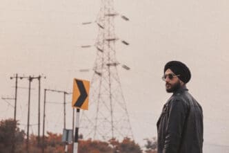 A man with a black turban, a beard, and sunglasses looks to the side while standing in front of a large electrical transmission tower under a hazy sky.