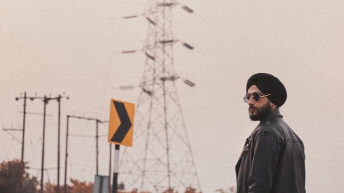 A man with a black turban, a beard, and sunglasses looks to the side while standing in front of a large electrical transmission tower under a hazy sky.