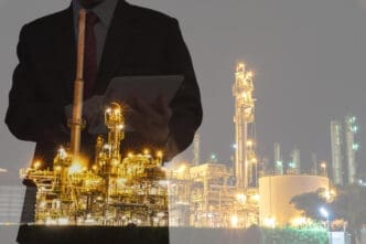 A double exposure image showing a person in a business suit holding a tablet, superimposed over a brightly lit industrial plant at night.