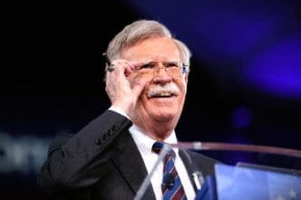 A close-up, professional photograph of John Bolton smiling and adjusting his glasses while speaking at a podium on a stage with a dark background.