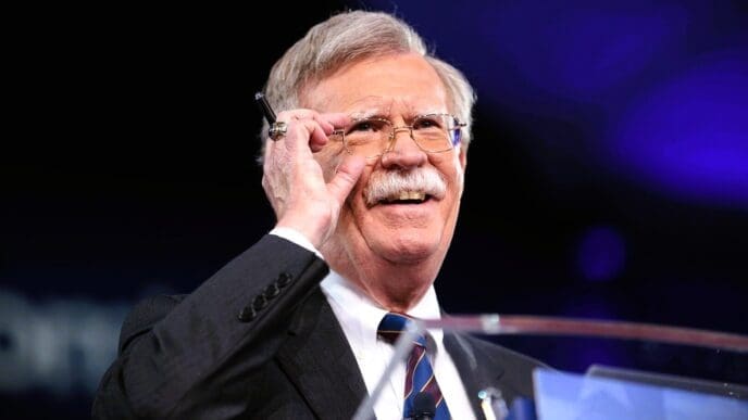 A close-up, professional photograph of John Bolton smiling and adjusting his glasses while speaking at a podium on a stage with a dark background.