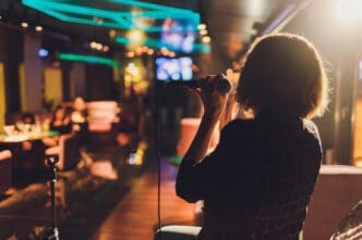 A person stands with their back to the camera, illuminated by a spotlight, singing into a microphone in a dimly lit bar with blurry patrons in the background.