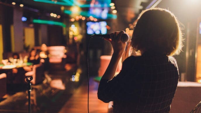 A person stands with their back to the camera, illuminated by a spotlight, singing into a microphone in a dimly lit bar with blurry patrons in the background.