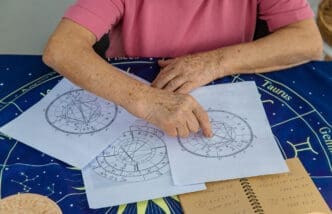 An astrologer's hands point to an intricate birth chart on a table, which is covered with a dark blue mat featuring zodiac signs, symbolizing an analysis of one's karmic blueprint in Vedic astrology.