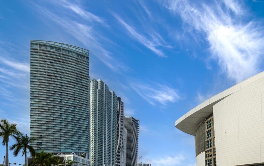A low-angle view of the Kaseya Center, a large white building with a sloping roof, next to several modern, curved skyscrapers under a bright blue sky with wispy clouds.