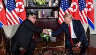 President Donald Trump and North Korean leader Kim Jong Un are seated and shaking hands in front of the flags of their respective nations.