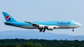 A large Korean Air Boeing 747-8 passenger jet, painted in a light blue and white livery, is seen in flight with its landing gear deployed.