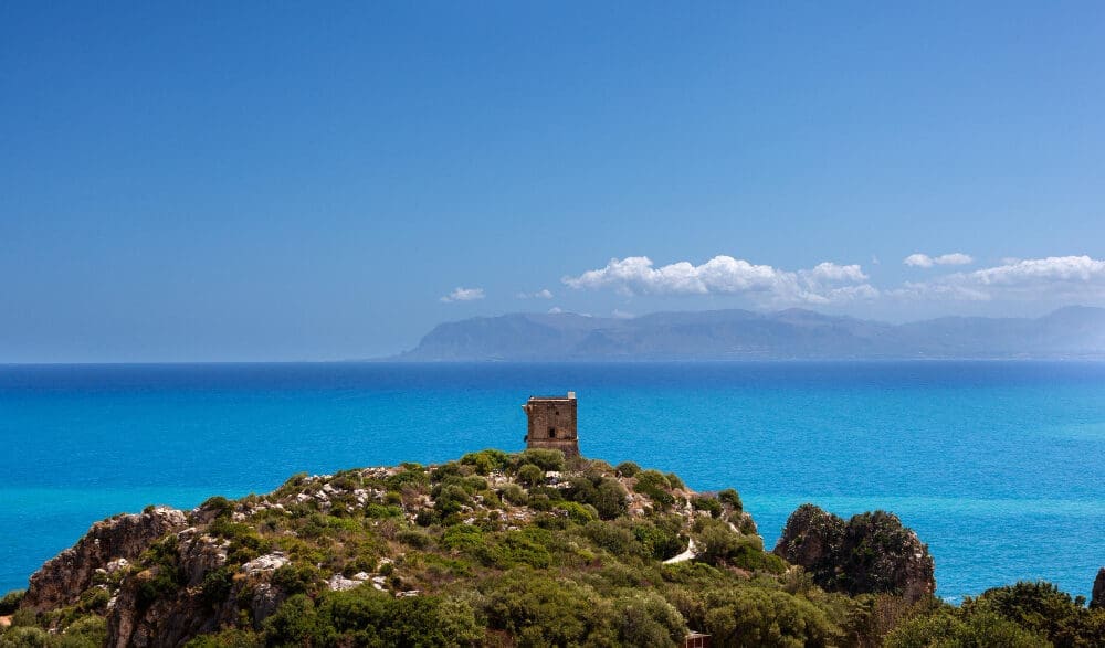 A small, ancient stone watchtower sits on a rocky, green hill overlooking the vast, turquoise Mediterranean Sea, with distant mountains on the horizon under a clear blue sky, in Scopello, Sicily.