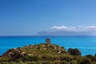 A small, ancient stone watchtower sits on a rocky, green hill overlooking the vast, turquoise Mediterranean Sea, with distant mountains on the horizon under a clear blue sky, in Scopello, Sicily.