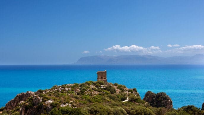 A small, ancient stone watchtower sits on a rocky, green hill overlooking the vast, turquoise Mediterranean Sea, with distant mountains on the horizon under a clear blue sky, in Scopello, Sicily.
