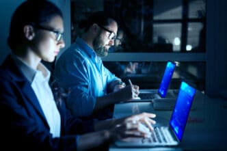 A man and a woman with glasses work late at night at a table, both focused on their laptops, with the man also writing in a notebook, in a dark, blue-lit office environment.