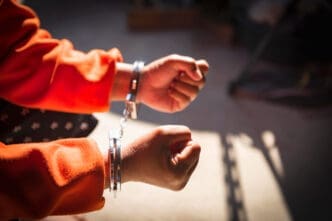 A close-up shot of a person's hands in handcuffs, wearing an orange jumpsuit.