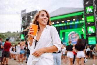 A happy young woman with long blonde hair, wearing a white shirt and black top, holds an orange cup and smiles while looking away from the camera at a music festival with a stage and a crowd in the background.
