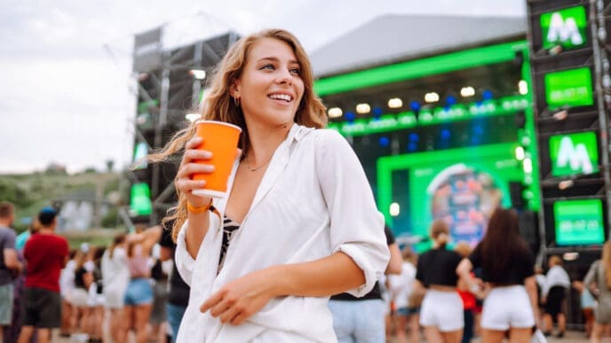 A happy young woman with long blonde hair, wearing a white shirt and black top, holds an orange cup and smiles while looking away from the camera at a music festival with a stage and a crowd in the background.