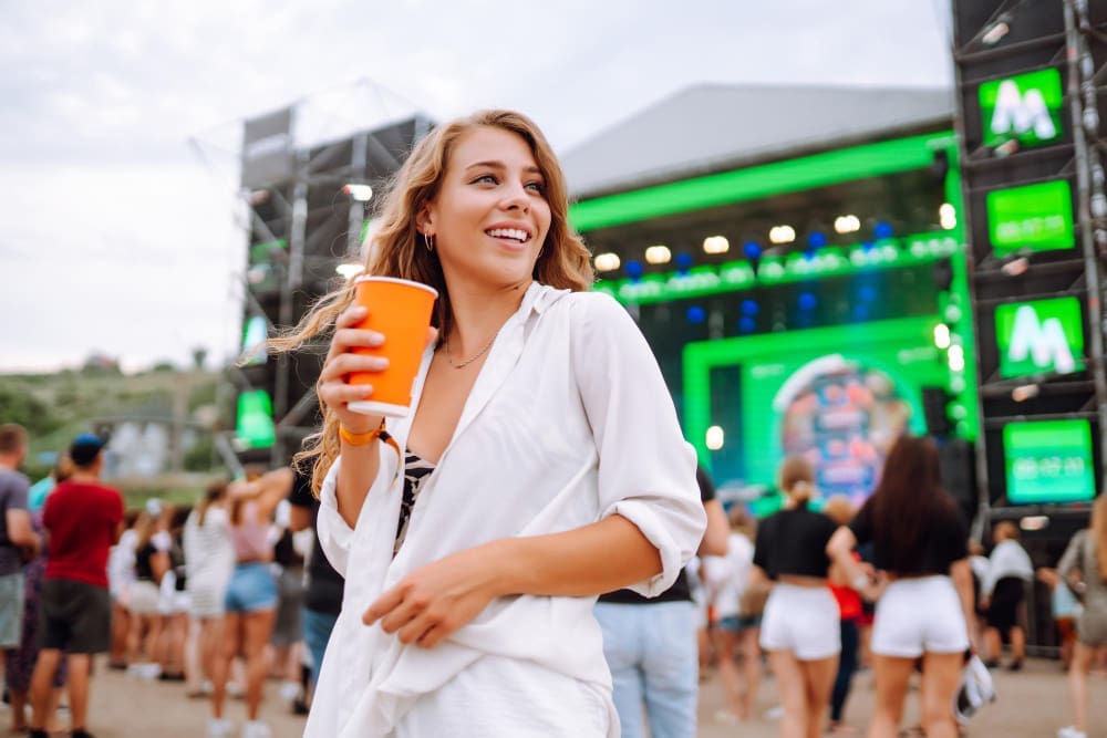 A happy young woman with long blonde hair, wearing a white shirt and black top, holds an orange cup and smiles while looking away from the camera at a music festival with a stage and a crowd in the background.