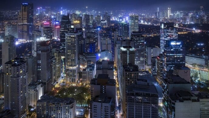 A panoramic night view of the dense urban skyline of Makati City, with all the skyscrapers and streets illuminated by countless lights.
