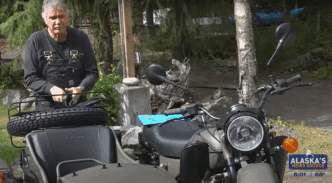A screenshot from a news report showing a man in overalls standing next to a motorcycle with a sidecar.