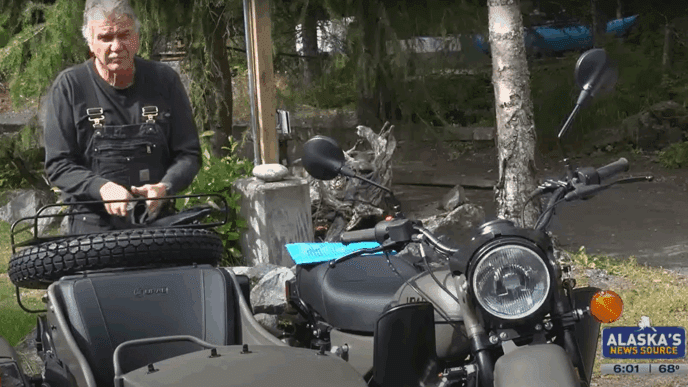 A screenshot from a news report showing a man in overalls standing next to a motorcycle with a sidecar.