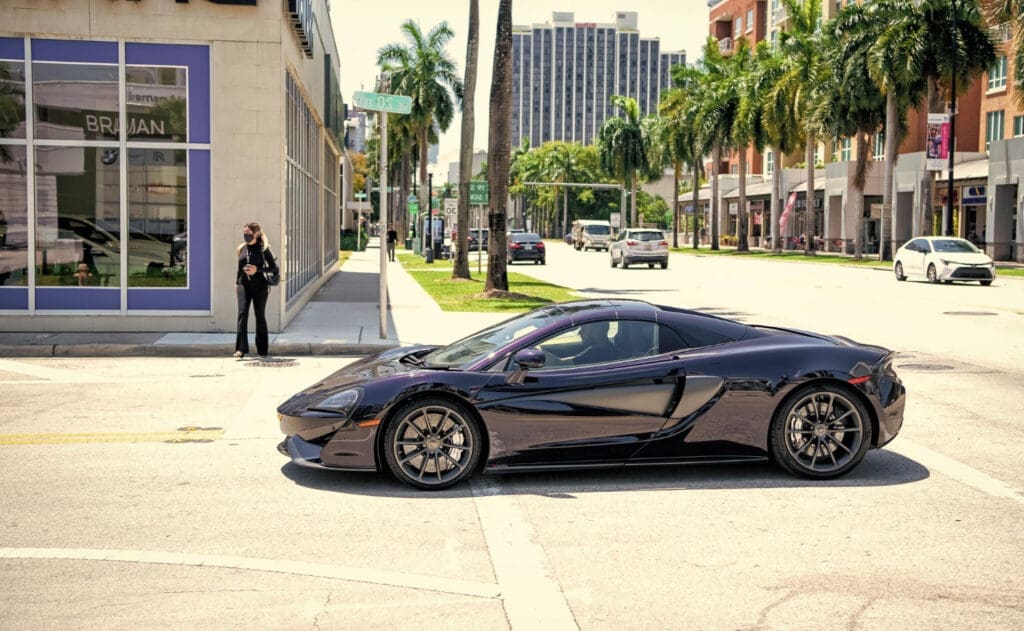 A side-view shot of a dark, sleek McLaren 570GT sports car driving on a street lined with modern buildings and palm trees in the Miami Design District.