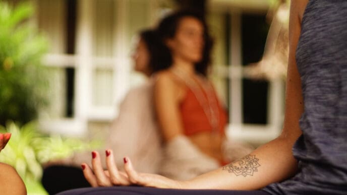 A close-up photograph of a woman's arm with a mandala tattoo, resting on her knee during a meditation or yoga session, with other people blurred in the background.