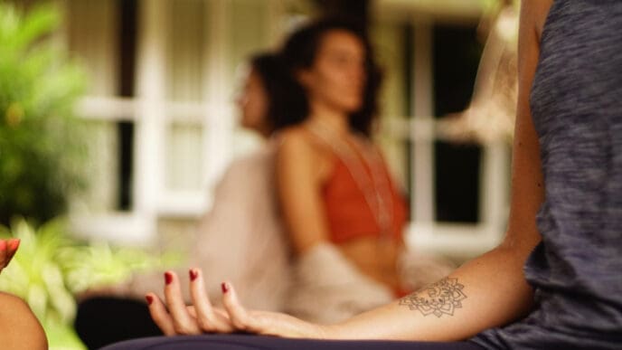 A close-up photograph of a woman's arm with a mandala tattoo, resting on her knee during a meditation or yoga session, with other people blurred in the background.