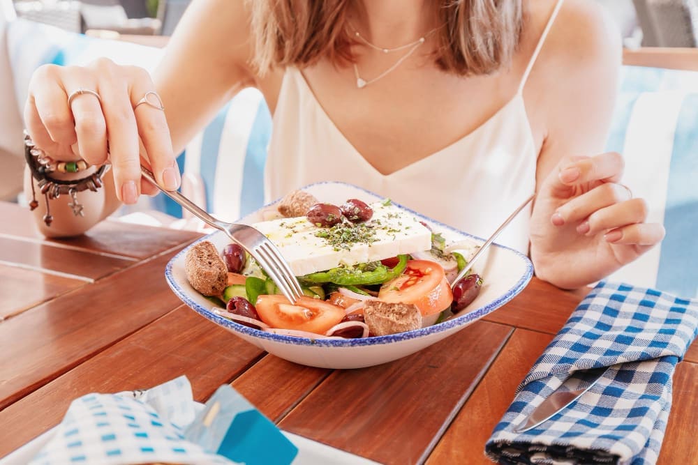 A person is about to eat a large bowl of Greek salad with a large block of feta cheese, tomatoes, and olives.