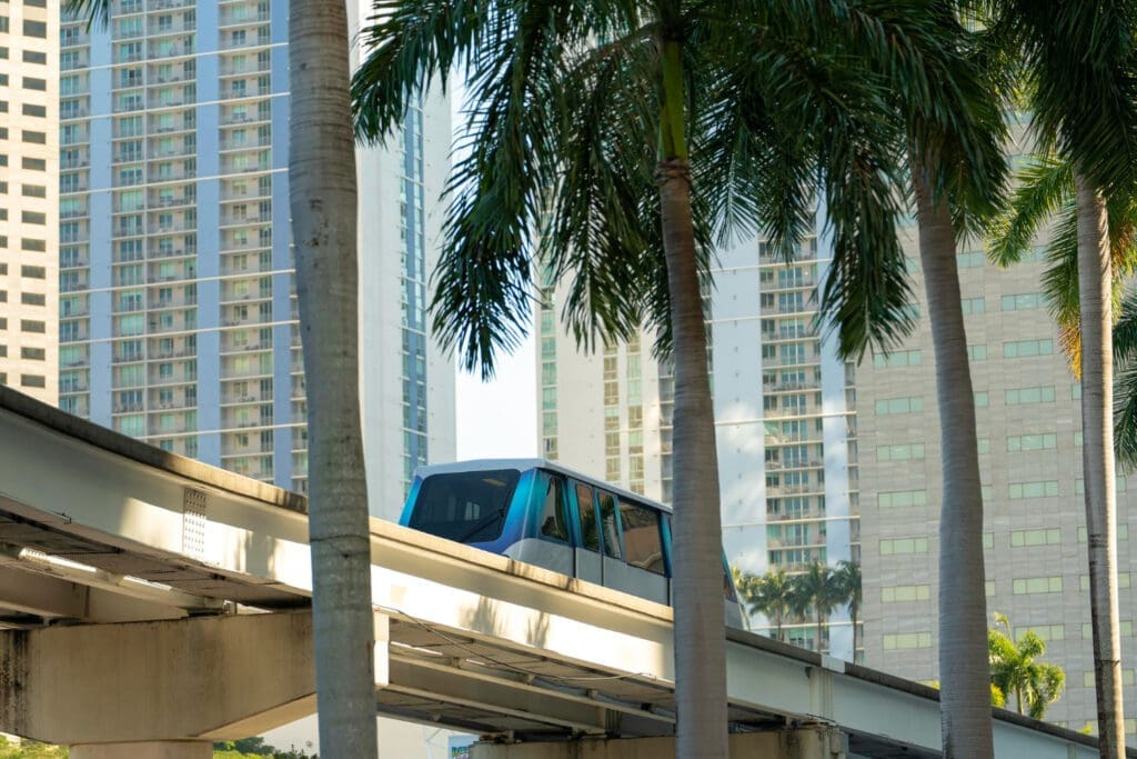 A Miami Metromover train moves on an elevated track among palm trees with high-rise buildings in the background