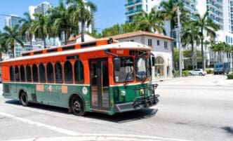 An orange and green Miami Trolley bus is driving down a street lined with palm trees and modern buildings, under a clear blue sky.