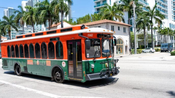 An orange and green Miami Trolley bus is driving down a street lined with palm trees and modern buildings, under a clear blue sky.