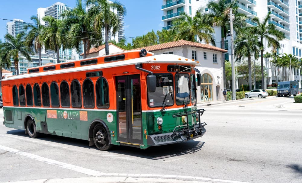 An orange and green Miami Trolley bus is driving down a street lined with palm trees and modern buildings, under a clear blue sky.
