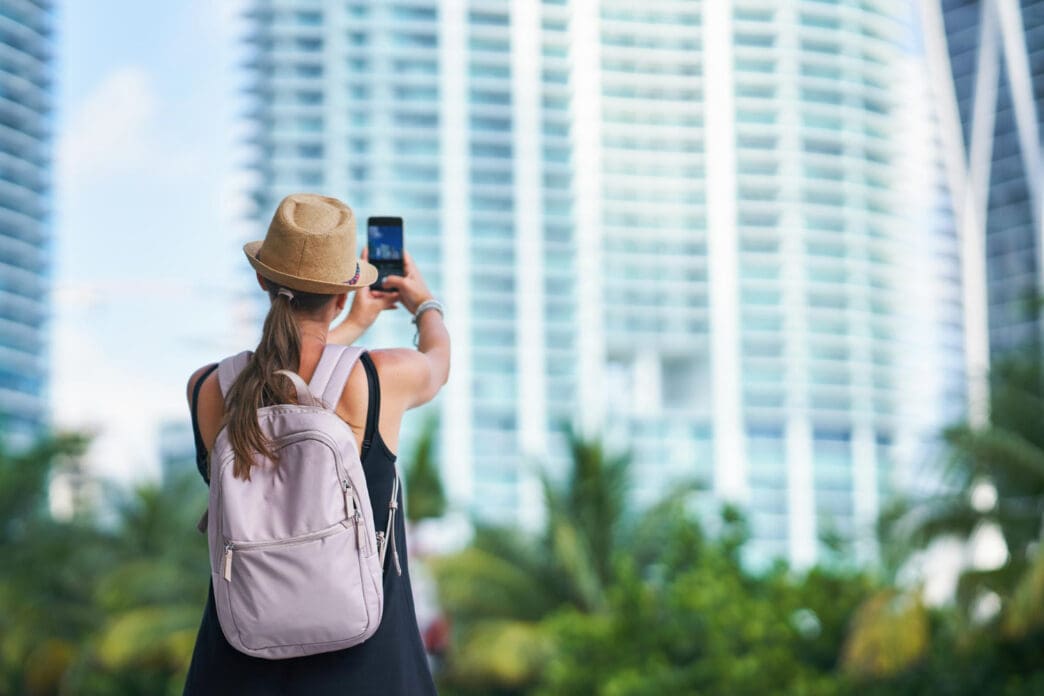 A woman wearing a sun hat and a backpack is taking a picture of a modern high-rise building in downtown Miami with her smartphone.