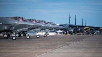 A line of several F-35 fighter jets is parked on a military tarmac, with a large B-52 bomber in the background.