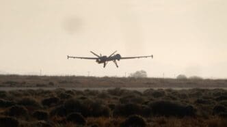 A large military drone, a Predator or Reaper type, is seen landing on a runway, silhouetted against a hazy sky with dry, bushy terrain in the foreground.
