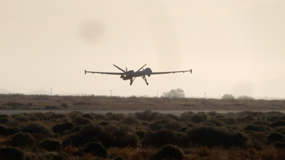 A large military drone, a Predator or Reaper type, is seen landing on a runway, silhouetted against a hazy sky with dry, bushy terrain in the foreground.