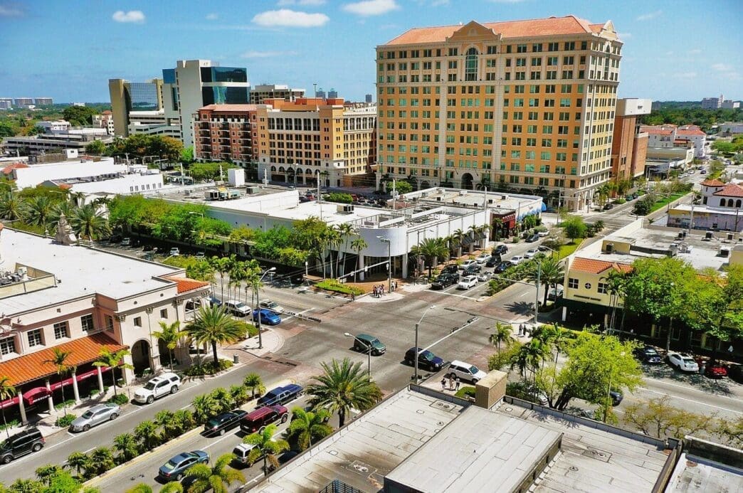 An aerial view of Miracle Mile in Coral Gables, Florida, showing a busy street intersection with Mediterranean-style buildings and palm trees.