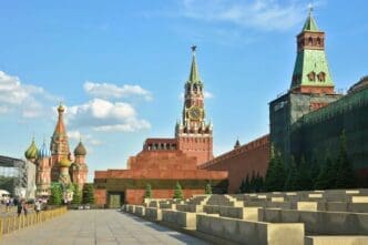 The Kremlin wall and Spasskaya Tower are visible, with the stepped Lenin Mausoleum in the foreground and the domes of St. Basil's Cathedral in the distance under a blue sky.