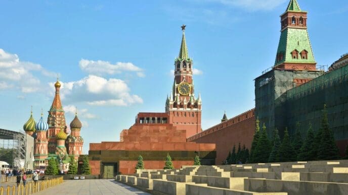 The Kremlin wall and Spasskaya Tower are visible, with the stepped Lenin Mausoleum in the foreground and the domes of St. Basil's Cathedral in the distance under a blue sky.
