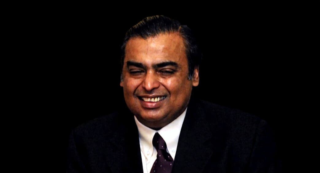 A close-up portrait of Mukesh Ambani, smiling and wearing a dark suit and tie.