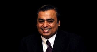 A close-up portrait of Mukesh Ambani, smiling and wearing a dark suit and tie.