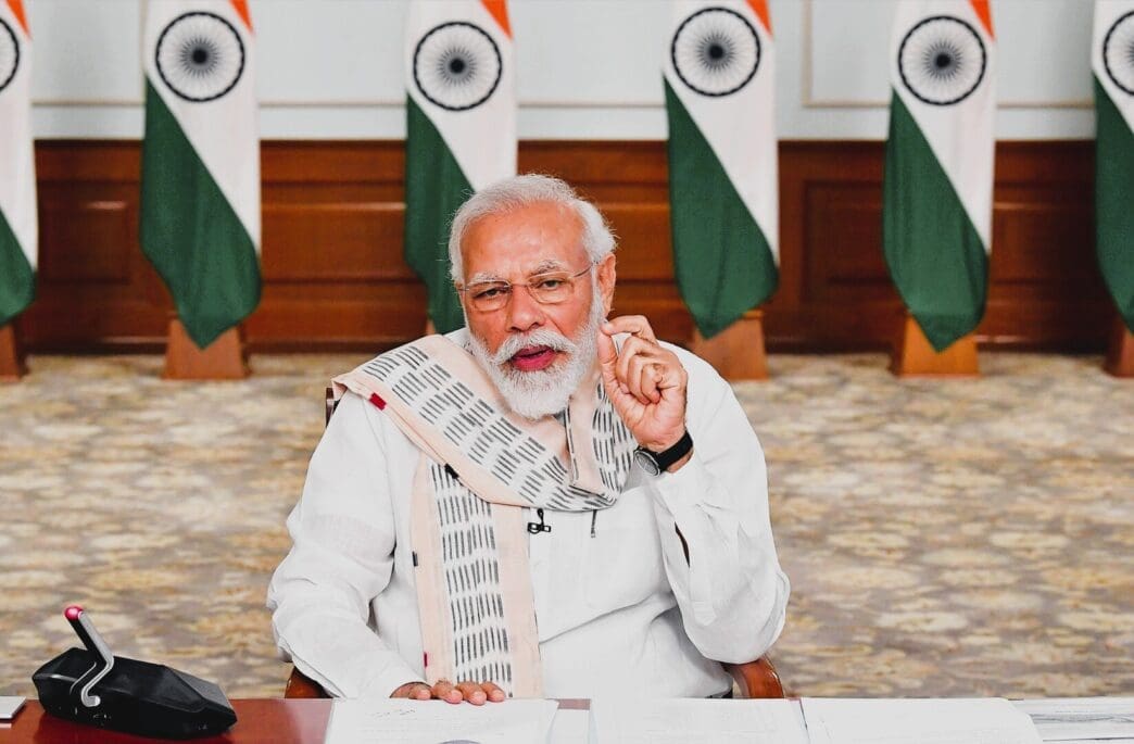 Indian Prime Minister Narendra Modi speaks while seated at a desk, with Indian flags in the background