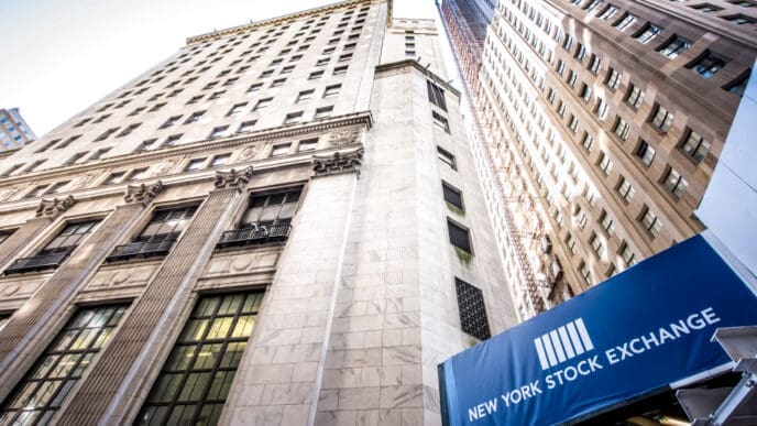 The New York Stock Exchange building, seen from a low angle, with a blue sign and a tall skyscraper in the background.