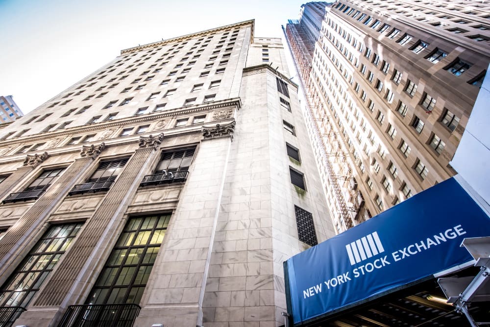 The New York Stock Exchange building, seen from a low angle, with a blue sign and a tall skyscraper in the background.