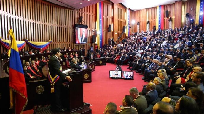 Nicolas Maduro, wearing a presidential sash, stands at a podium in a large, red-carpeted chamber with a seated audience, as he is sworn in as president.