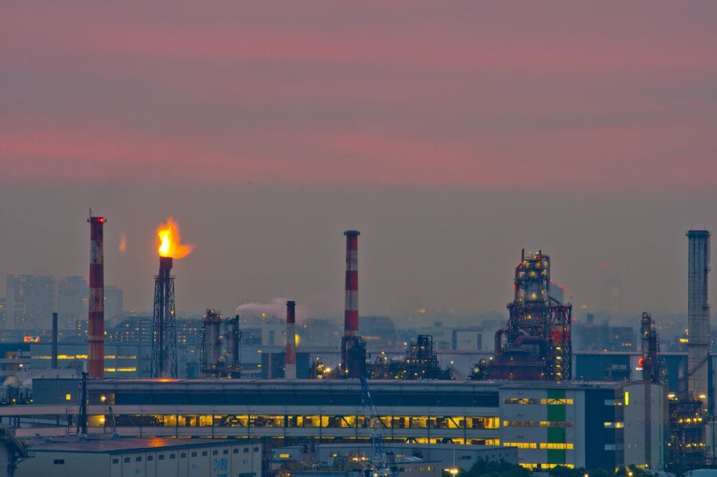 A Russian oil refinery with multiple smokestacks and a large flare stack burning against a hazy, pink-and-purple sunset sky